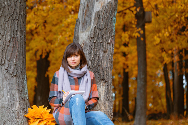 University Park, studie concept. Portret van een jonge vrouw in herfst bos. Schrijft in een notitieblok. Brunette vrouw in herfst park met modieuze plaid jas en sjaal. - Foto, afbeelding