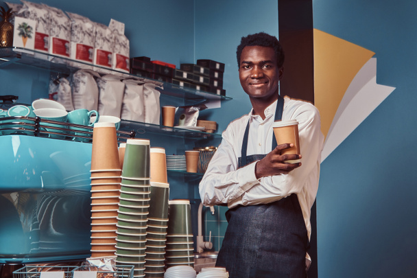 Handsome African American barista in uniform holds a cup of coffee while standing in her coffee shop. - Foto, Bild