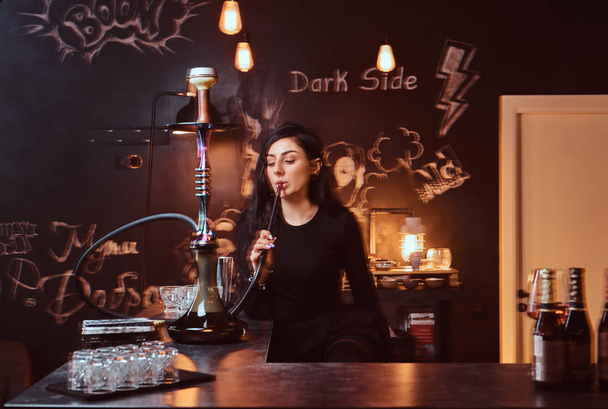Sexy brunette meisje in verleidelijke zwarte kleding rookt een waterpijp zittend op de teller in een nachtclub. - Foto, afbeelding