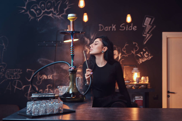 Hot brunette girl in seductive black clothes smokes a hookah while sitting on the counter in a nightclub. - Photo, Image