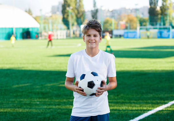 cute young boy in white blue sportswear holds classical black and white football ball on the green grass of the stadium field. Soccer game, training, hobby concept. with copy space - Foto, immagini