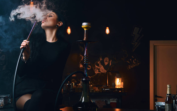 sexy brünettes Mädchen in verführerischer schwarzer Kleidung raucht eine Wasserpfeife, während es in einem Nachtclub oder einer Bar auf dem Tresen sitzt. - Foto, Bild