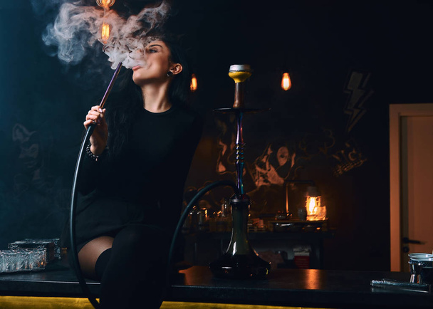 Το σέξι κορίτσι μελαχρινή στο σαγηνευτικό μαύρα ρούχα καπνίζει ναργιλέ ενώ κάθεται στον πάγκο σε ένα νυχτερινό κέντρο διασκέδασης ή το μπαρ. - Φωτογραφία, εικόνα