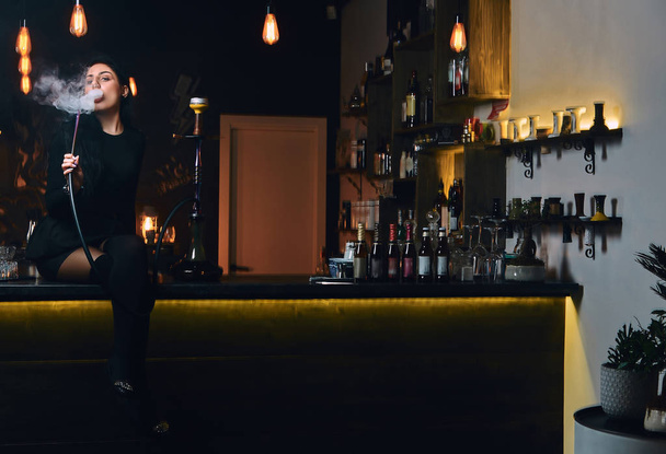 Menina morena sexy em roupas pretas sedutoras fuma um narguilé enquanto está sentado no balcão em uma boate ou bar
. - Foto, Imagem
