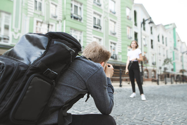 Professionele fotograaf in een grijs shirt en een rugzak gefotografeerd een meisje op de straten van de stad. Focus op de fotograaf. Professionele fotograaf neemt een fotosessie. - Foto, afbeelding
