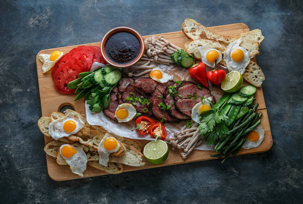 Breakfast meals variety flat lay. Top view on buffet wooden board with meat, eggs, vegetables, bread etc - Photo, Image