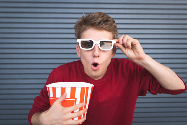Patlamış mısır onun elinde bir fincan ile 3d gözlük bir film izlemek şaşırmış bir genç adam portresi. Duygusal öğrenci bir film sinemada izlerken. Koyu arka plan. Boşaltmak - Fotoğraf, Görsel