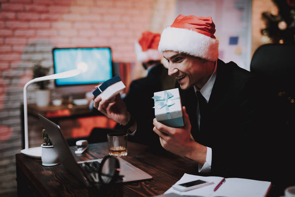 Businessman in Red Cap in Office on New Year Eve. Christmas Tree in Office. Laptop on Desk. Business Concept. Man in Black Suit. Celebrating of New Year. Using Digital Device. Gift Box. - Fotoğraf, Görsel