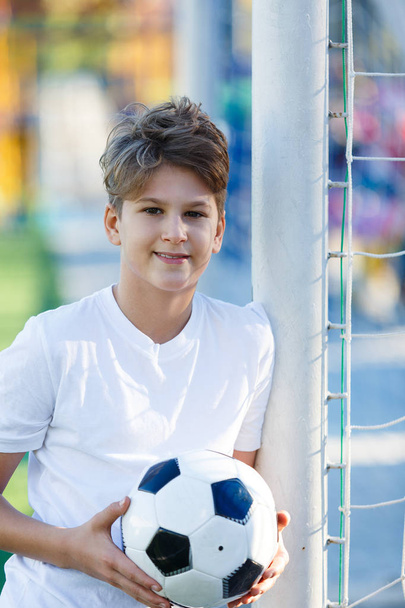 cute young boy in white blue sportswear holds classical black and white football ball on the green grass of the stadium field. Soccer game, training, hobby concept. with copy space  - Foto, afbeelding