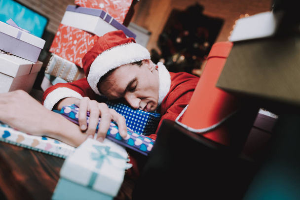 Young Man in Santa Claus Costume with Gift Boxes. New Year Eve. Christmas Tree in Office. Celebrating of New Year. Red Costume. Man in Red Cap. Pile of Boxes. Man Sleeping on Table. - Photo, image