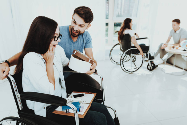 People in Wheelchair. Disabled in Hall. Woman in Wheelchair. Man on Crutches. Surprised Woman in Wheelchair. Room with Panoramic View. Gray Sofa. White Interior. Limited Opportunities. Gives Gift Box. - Fotoğraf, Görsel