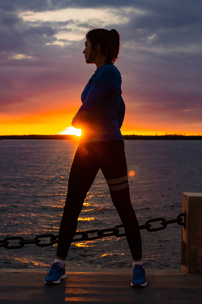 An attractive woman in sports clothes doing sports exercises in nature against the sunset and the Amur river, loves gymnastics, kneads her legs. Active young girl engaged in sports, leads a healthy lifestyle. - Photo, Image