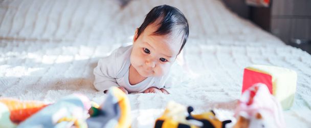 Lindo bebé regordete con una sonrisa feliz acostado en su estómago en una cama con una fila de juguetes suaves detrás de una pared de ladrillo
. - Foto, imagen