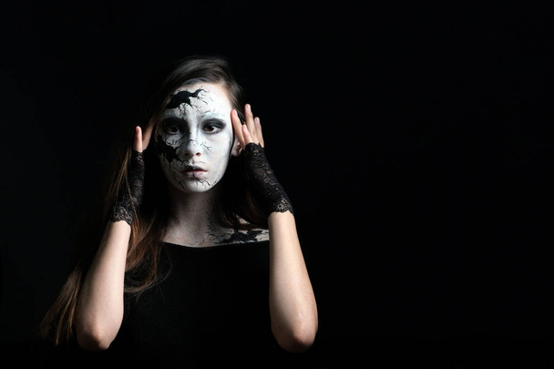 Makeup in the style of Halloween. A young beautiful girl with painted cracks on her face holds fingers of her temples near her eyes. Black background. Copy space. Dark dress and openwork gloves. The image of a witch or cracked doll. - Fotografie, Obrázek