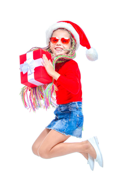 Girl in Santas hat. Portrait of little cute girl holding box of Christmas present, Small girl hug her gift with happy and fun expression isolated on white background. Jump up. Holiday gift - Foto, Imagen