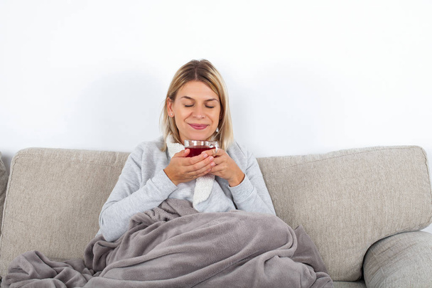 Blonde attractive woman drinking hot tea while sitting on the couch, wrapped in a grey blanket - Foto, Imagem