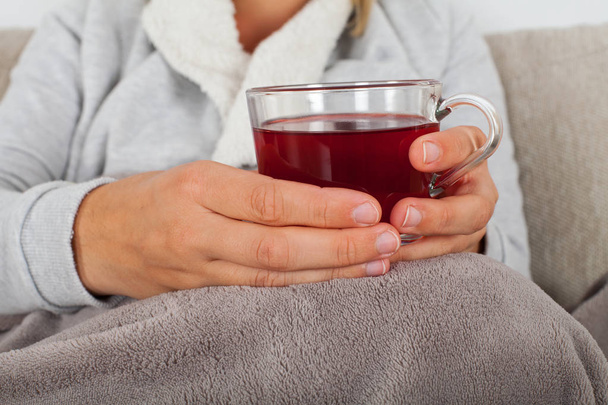 Close up picture of sick woman holding a cup of hot tea while sitting on the couch wrapped in a warm blanket - Фото, изображение