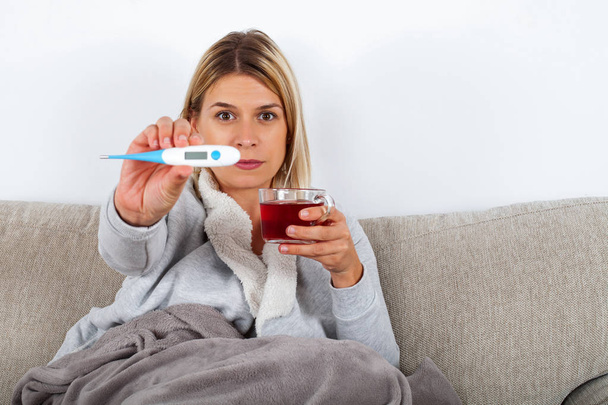 Sick woman sitting on the couch wrapped in a warm blanket, holding a hot tea and thermometer - Photo, Image
