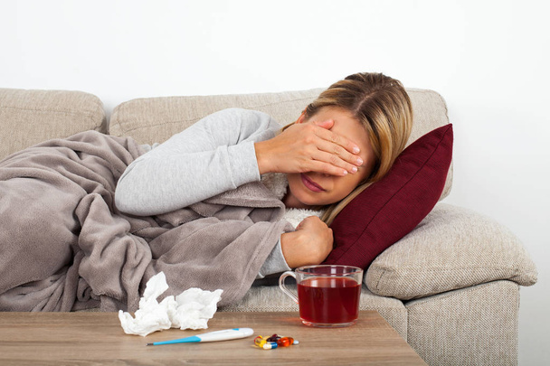Больная женщина лежит на диване, завернутая в теплое одеяло - лекарства, термометр и чай на столе
 - Фото, изображение