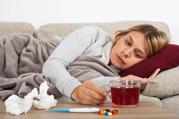 Sick woman lying on the couch wrapped in a warm blanket - medication, thermometer and tea on the table - Foto, Imagem