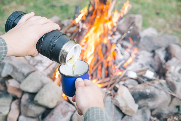 男は、金属のカップに紅茶を注ぐ。背景にキャンプの火。ハイキング - 写真・画像