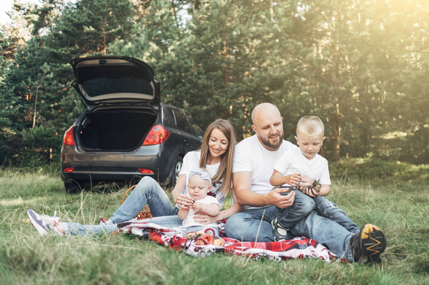 Mladý pár s jejich malé děti mají piknik v lese, pojem rodinný víkend, čtyři lidé se těší letní - Fotografie, Obrázek