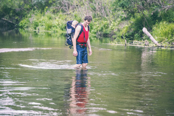 Ein Mann mit Kind überquert den Fluss. Der Vater trägt den Sohn im Rucksack. Rettung vor Überschwemmungen. Evakuierung der Flutopfer in Asien. Reisen mit einem Kind durch den Dschungel. Extremtourismus. - Foto, Bild