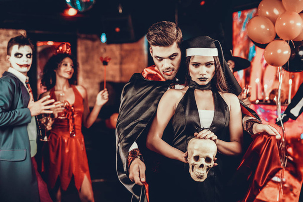 Portrait of Young Couple in Halloween Costumes. Beautiful Woman and Handsome Young Man Wearing Costumes holding Skull at Halloween Party in Nightclub. Happy Friends having Fun Celebrating Halloween - Foto, afbeelding