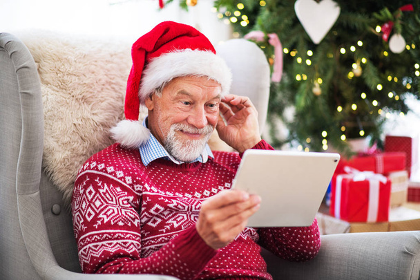 Ένα πορτρέτο του ανώτερος άνθρωπος με tablet που κάθεται στην πολυθρόνα στο σπίτι στο χρόνο Χριστουγέννων. - Φωτογραφία, εικόνα