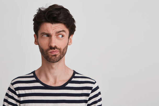 Clouse up of young brunet with beard looking to the right with distrust and discontent, frowning brows, lips pursed wears black and white striped tshirt, stands leftside isolated over white background - Photo, Image