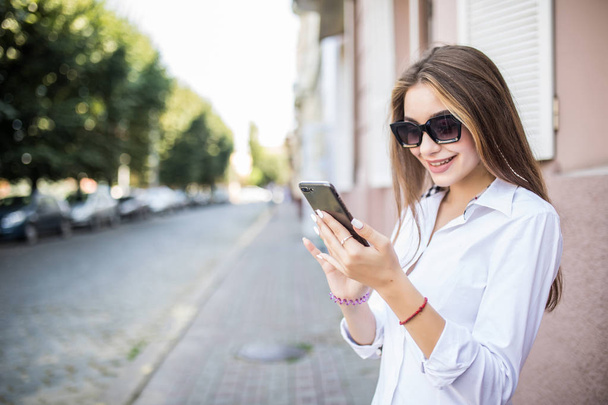 Mujer feliz sonriendo y caminando en la calle usando un teléfono inteligente y mirando a la cámara - Foto, imagen