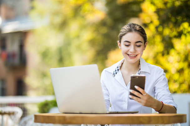 Mladá podnikatelka sedí u stolu v kavárně venku a využití smartphone na stůl šálek kávy a notebook - Fotografie, Obrázek