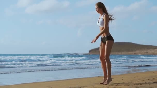 Dívka v sportovní kraťasy a tričko provádí skoky s dřepy na pláži nedaleko oceánu na Kanárských ostrovech. Zdravého životního stylu a fitness na dovolené. Krásné a zdravé tělo - Záběry, video