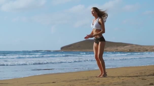Dziewczyna w sprawny spodenki i Koszulka wykonuje skoki z kuca i klaszcze na plaży, w pobliżu oceanu na Wyspach Kanaryjskich. Zdrowy styl życia i fitness na wakacje. Piękne i zdrowe ciało - Materiał filmowy, wideo