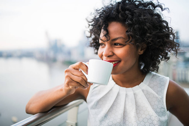 A portrait of a woman standing on a terrace, drinking coffee. - 写真・画像