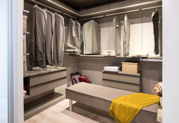 Interior of a walk in closet with hanging clothes and built in drawer units and seating in a luxury modern home - Photo, Image