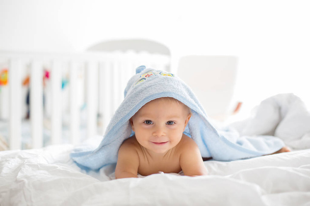 Little cute baby boy, lying in bed with towel after bath, sunny bedroom, white sheets - Photo, Image