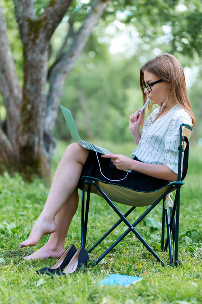 Young woman with a laptop sitting in the Park on a folding chair and holding a pen in her hand - Fotoğraf, Görsel