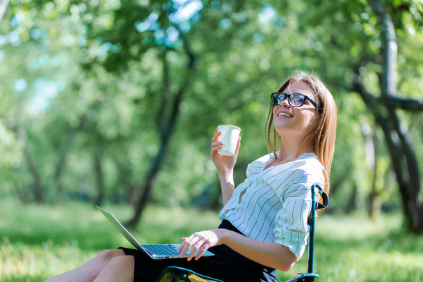 Girl using laptop on lunch break in city park and hold coffe in her hand - Fotó, kép