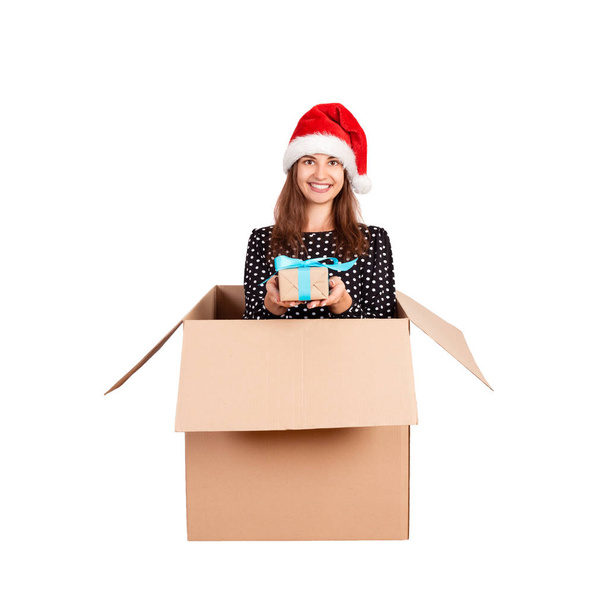 Joyeux femme heureuse en robe et chapeau de Noël s'amuser tout en étant assis dans la grande boîte-cadeau et en donnant cadeau ruban bleu witj. isolé sur fond blanc
. - Photo, image