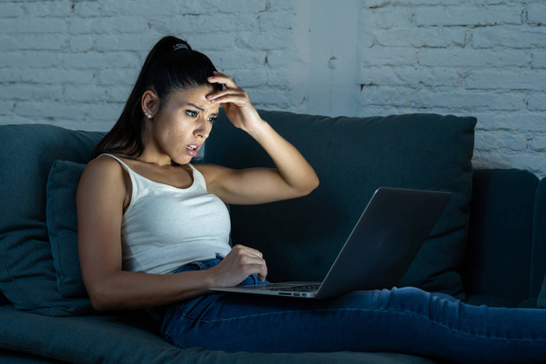 Beautiful young latin woman surfing on the internet on her computer laptop on the sofa at home late at night in new technology internet addiction and workaholic concept. - Photo, image