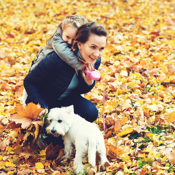 Madre e hija en otoño pasean con el perro. Feliz familia amorosa divirtiéndose. Pequeño perro blanco y familia disfrutando juntos al aire libre. Concepto de familia, mascotas y estilo de vida. Vacaciones de otoño
. - Foto, Imagen