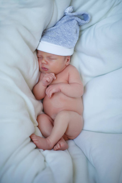 Newborn baby boy in a blue cap lies on a white bedspread and sleeps. The concept of a happy childhood, motherhood, breastfeeding. - Fotoğraf, Görsel