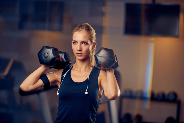 Mujer rubia levantando pesas mientras está de pie en un gimnasio. concepto de estilo de vida saludable
. - Foto, Imagen