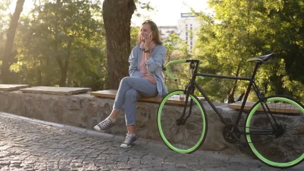 Fair haired girl sitting on the bench or parapet in the city park with paved her trekking bike next to her. Talking by her mobile phone. Wearing bright pink and blue coloured casual clothes and - Video
