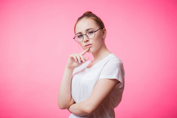 Portrait of a beautiful charming red haired woman with glasses on a pink background. Young girl wearing white t-shirt - Photo, image