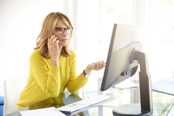 Shot of middle aged investment businesswoman using her cell phone and talking with client while sitting in front of laptop in the office.  - Photo, image