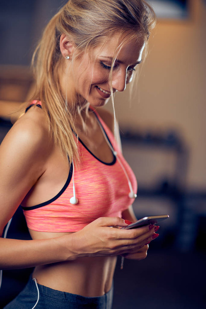 Young Caucasian woman using smart phone while standing in gym.  - Photo, image