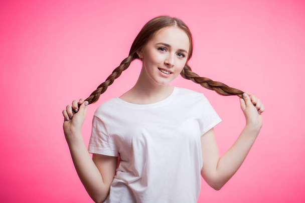 Young attractive girl holding for long braids. Portrait of beautiful smiling woman with beauty hair braid on pink background. Human emotions, facial expression concept - Photo, image