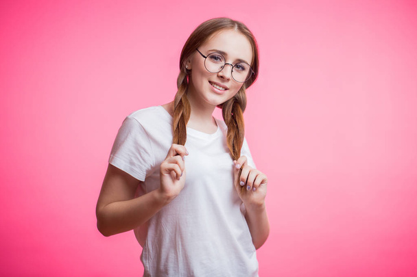 Happy young woman in glasses and white t-shirt looking at camera and smiling. Attractive woman holding hands with her pigtails, braid. Human emotions, facial expression concept - Photo, Image